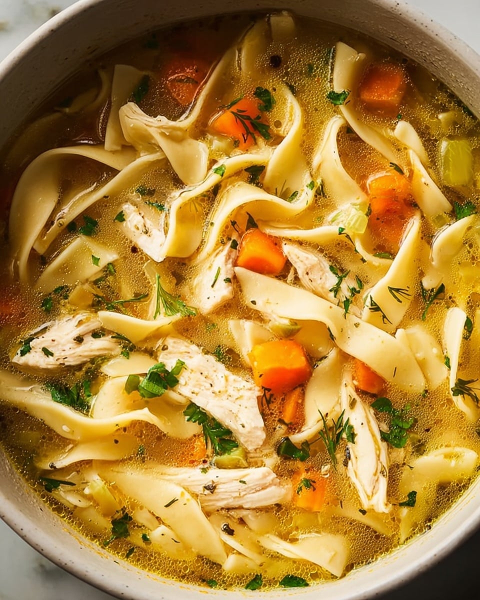 A close-up view inside a black pot filled with chicken noodle soup showing three layers: the bottom layer is clear golden broth, the middle layer has wide, flat pale yellow noodles twisted and floating, and the top layer features bright orange carrot slices, white shredded chicken pieces, and fresh chopped green herbs scattered evenly on the surface. A silver spoon peeks in from the left side, partially submerged in the soup. The pot is set on a white marbled surface, with a blurred corner of a white bowl with chopped herbs visible in the background. Photo taken with an iphone --ar 4:5 --v 7