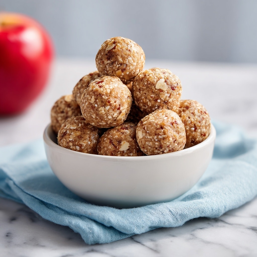 A white bowl filled with multiple round energy balls stacked closely together, each ball has a rough texture with visible bits of oats and a mix of light brown and golden colors, reflecting a chewy and slightly sticky surface. The bowl is placed on a soft teal cloth, and in the background, a red apple is slightly blurred. The scene is set on a white marbled surface. Photo taken with an iphone --ar 4:5 --v 7