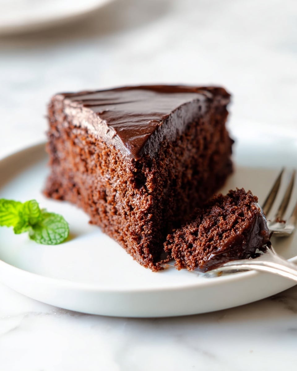 A close-up view of a single slice of chocolate cake resting on a white plate set on a white marbled surface. The cake has two layers: a moist dark brown chocolate sponge base with visible crumbs and textured bits, topped with a thick, smooth layer of darker chocolate frosting with swirled, creamy peaks. A small green mint leaf sits beside the cake slice, adding a touch of color to the scene. Photo taken with an iphone --ar 4:5 --v 7