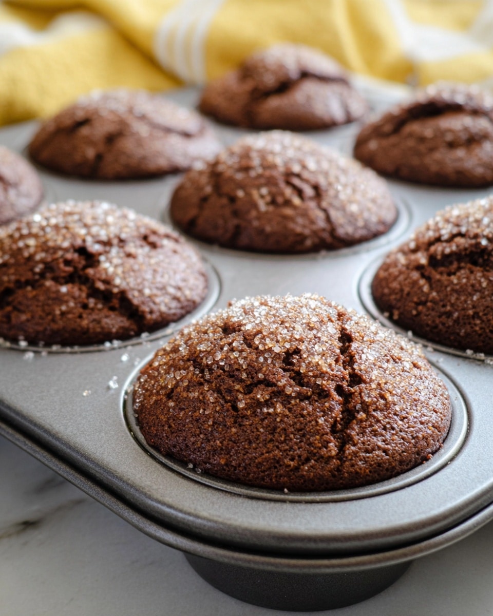 This image shows a close-up of several round chocolate muffins in a metal baking tray. Each muffin has a cracked top with a dark brown, moist texture and is covered with sparkling coarse sugar crystals, giving a crunchy appearance. The muffins are evenly spaced in the tray, resting on a white marbled surface. The overall look is warm and freshly baked. photo taken with an iphone --ar 4:5 --v 7