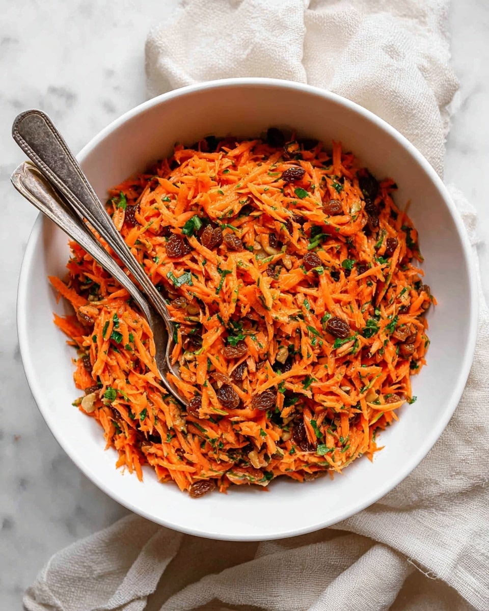 A white bowl sits on a white marbled surface, filled with a bright orange shredded carrot salad mixed with small brown raisins and green leafy herbs, sprinkled with light brown nut pieces. Two silver spoons rest inside the bowl, one slightly buried in the salad showing the texture of the grated carrots. A beige cloth partially covers the top right edge of the bowl, adding soft texture to the scene. photo taken with an iphone --ar 4:5 --v 7
