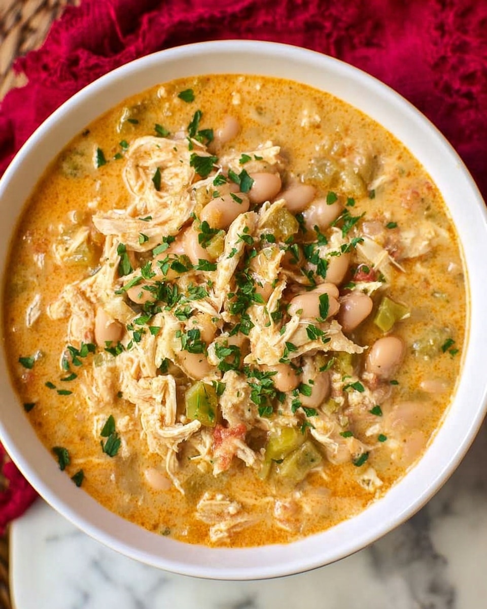 A white bowl filled with creamy white chicken chili sits at the center. The chili has a thick texture with chunks of white chicken, white beans, and small pieces of green pepper, all mixed in a light yellow creamy broth. The surface is sprinkled with black pepper and herbs, adding a touch of green. Behind the bowl, there is a small glass bowl with white shredded cheese and a white plate holding golden toasted bread sticks. The background is a white marbled surface with a red patterned cloth underneath the bowl. photo taken with an iphone --ar 4:5 --v 7