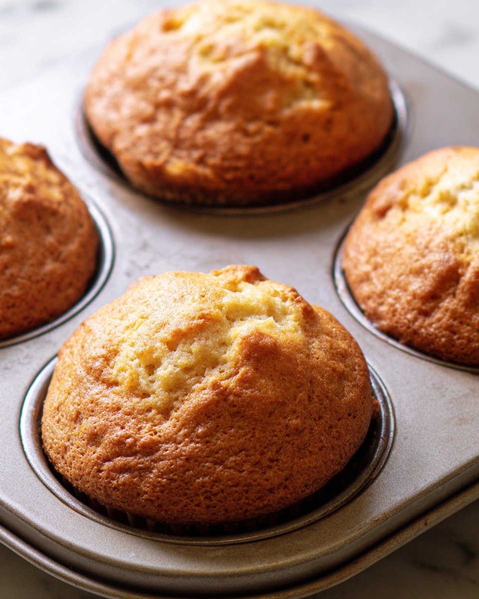 A close-up image of a golden brown muffin in a muffin tin, showing a rough, slightly cracked top with a soft, moist texture beneath the crust. The muffin is rounded and domed, filling its tin cup fully. Around it, two more muffins are partially visible in the metal muffin tray, all set on a white marbled surface with a cooling rack underneath. The overall look is warm and freshly baked, with soft natural light highlighting the muffin’s texture. Photo taken with an iphone --ar 4:5 --v 7