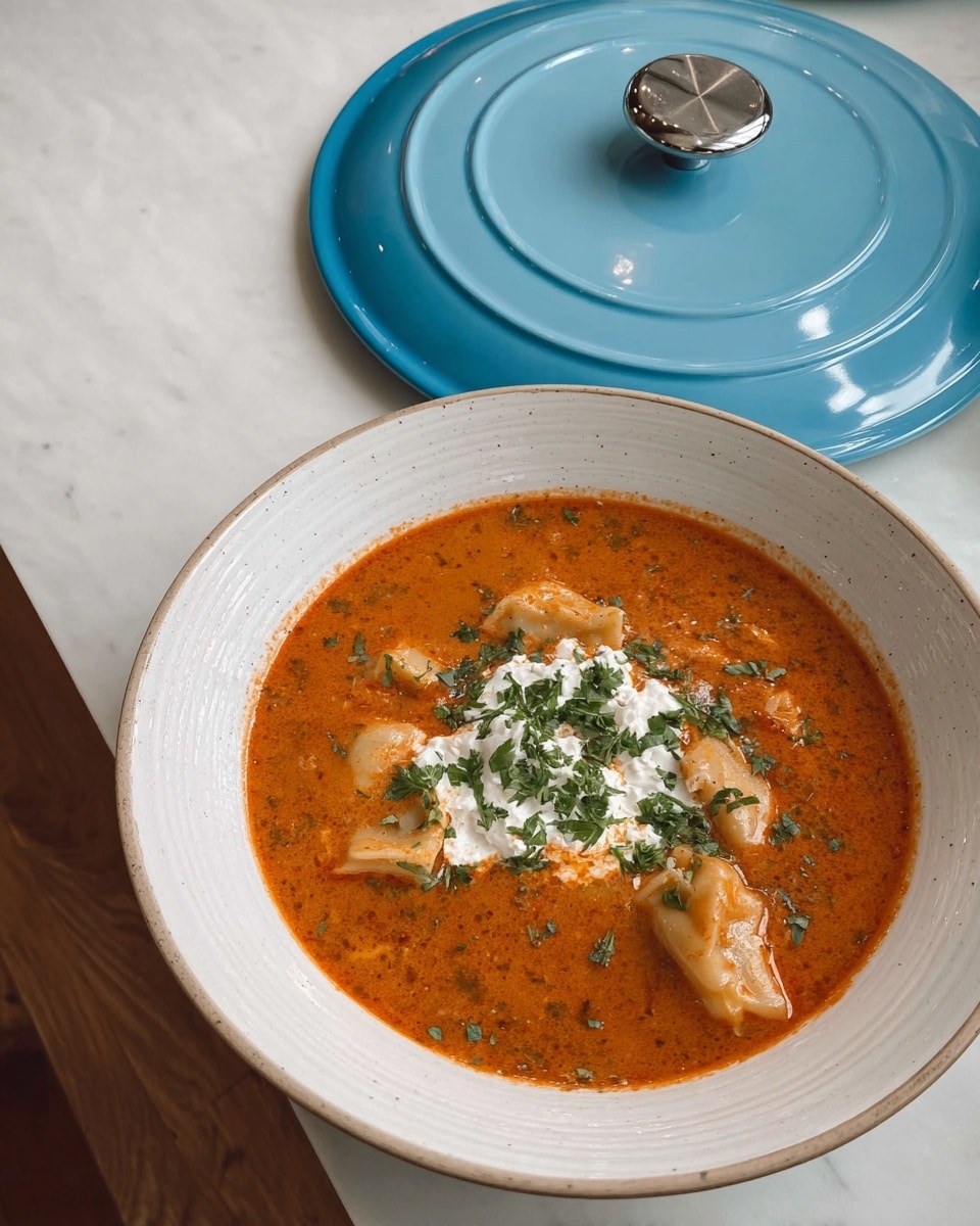 A white bowl filled with a rich, orange-red soup with visible black specks of herbs and spices. The soup contains several folded pasta pieces submerged in the liquid. On top, there is a dollop of white creamy cheese and green chopped herbs scattered around it for garnish. The bowl is placed on a wooden table next to a light blue pot lid with a silver handle. The background has a white marbled texture. photo taken with an iphone --ar 4:5 --v 7