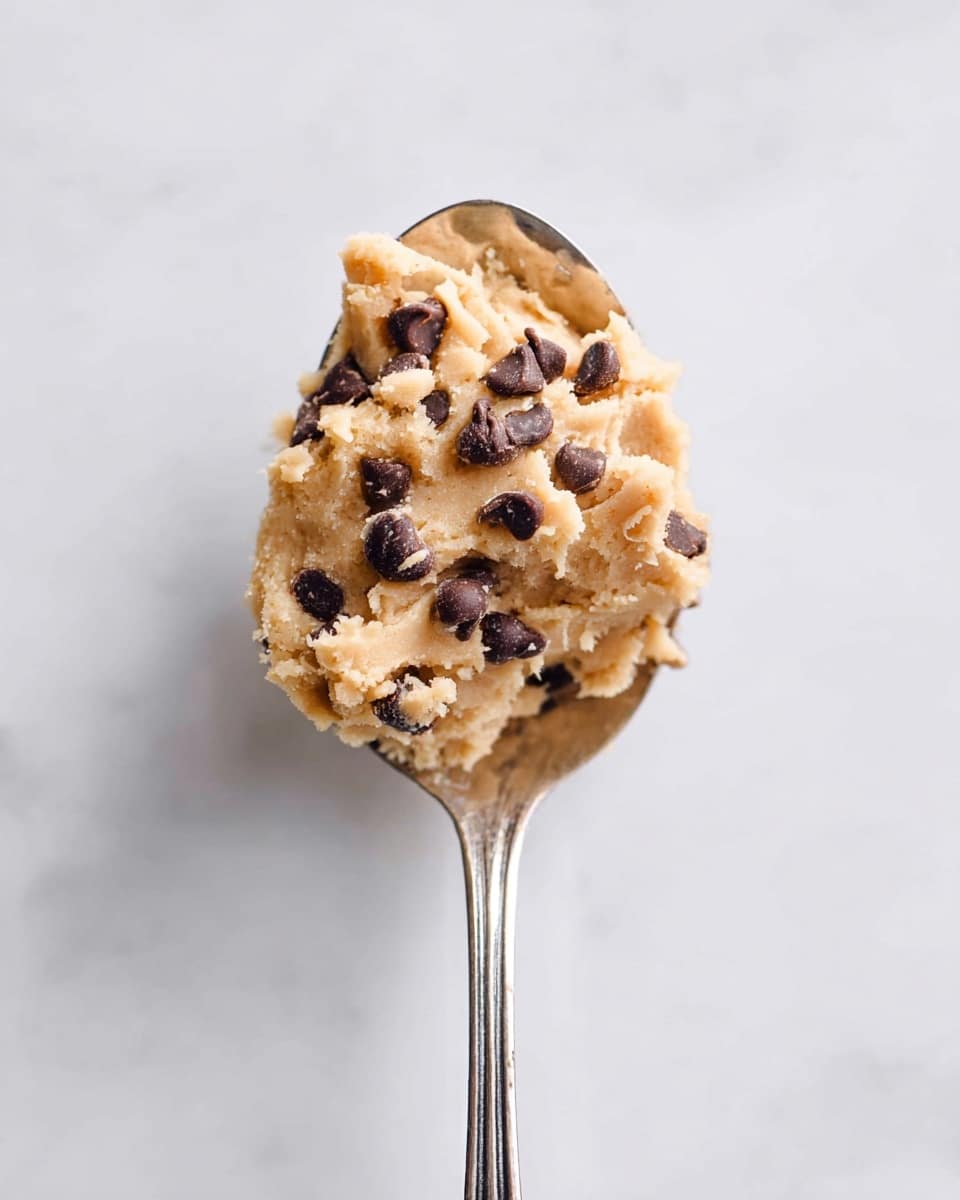 A close-up view of a silver spoon holding a scoop of light brown cookie dough mixed with small dark brown chocolate chips scattered throughout. The dough has a soft, slightly crumbly texture with a few chocolate chips poking out. The background shows a clean, white marbled surface that is smooth and bright, making the dough and spoon stand out clearly. photo taken with an iphone --ar 4:5 --v 7