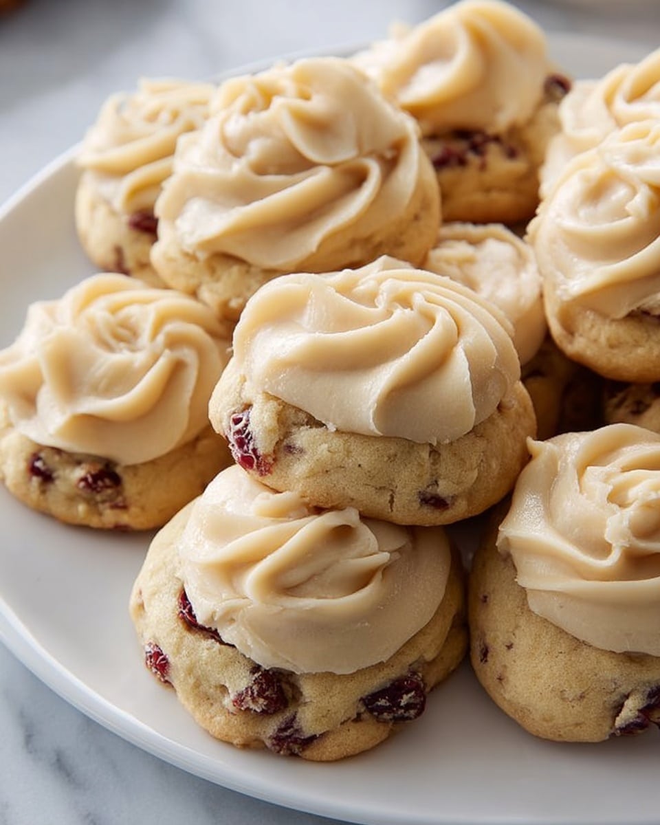 A white plate filled with a pile of small, round cookies that have a light golden-brown color with visible red cranberry pieces baked into them; each cookie is topped with a thick swirl of creamy, light beige frosting that has a smooth, slightly textured appearance. The cookies have a rough, slightly crumbly surface underneath the frosting. The setting is on a white marbled surface. photo taken with an iphone --ar 4:5 --v 7