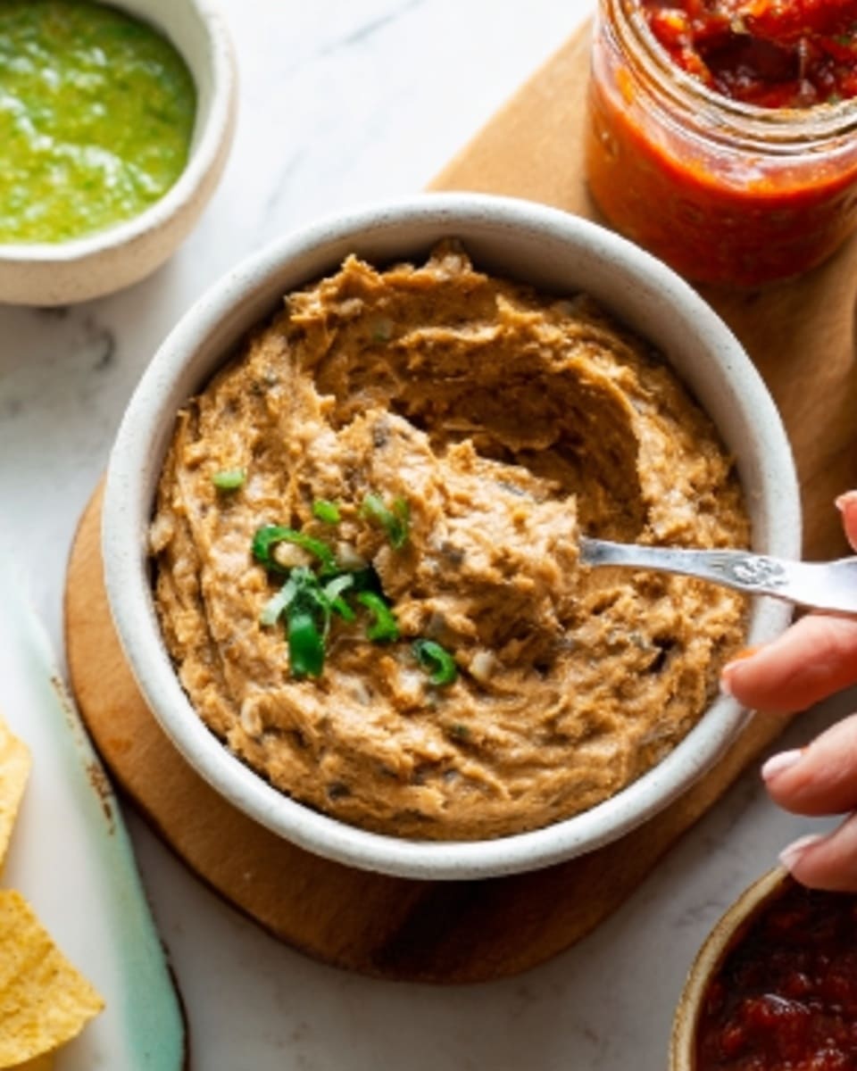 A white bowl filled with smooth, light brown refried beans topped with small green cilantro leaves sits on a wooden surface. A silver spoon is inside the bowl, slightly digging into the beans. Around the bowl, there are parts of other bowls visible: one white bowl with green guacamole, another white bowl with red salsa, and a small wooden bowl with light green sauce. The background is a white marbled texture. Photo taken with an iphone --ar 4:5 --v 7