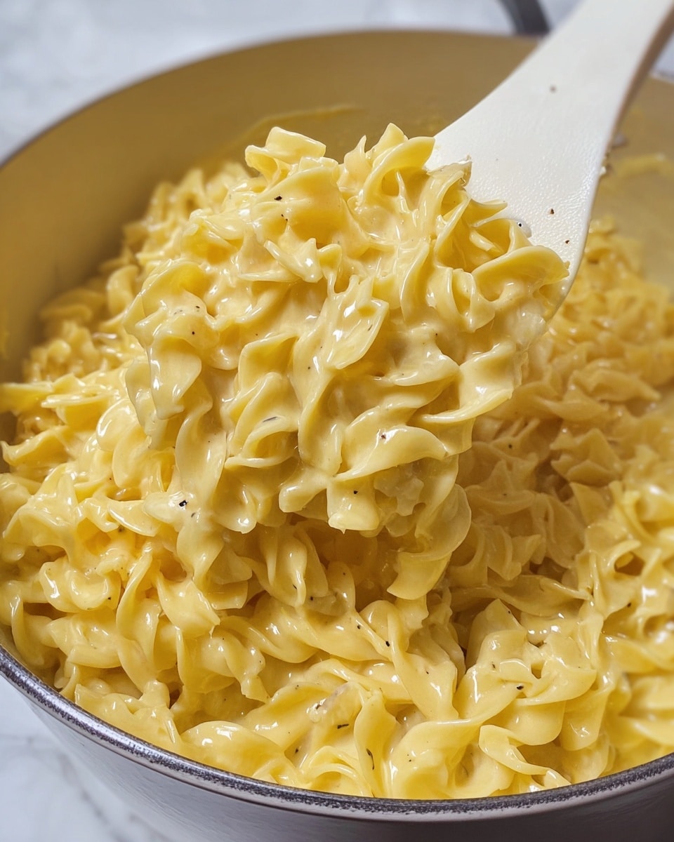 A large white pot filled with cooked egg noodles that are yellow and soft with a slightly glossy, creamy coating, showing some small black pepper specks throughout; the noodles fill the pot up to about four-fifths and have a wavy, curly texture with some overlapping layers. Behind the pot, a carton of chicken broth is partially visible, resting on a white marbled surface. Photo taken with an iphone --ar 4:5 --v 7