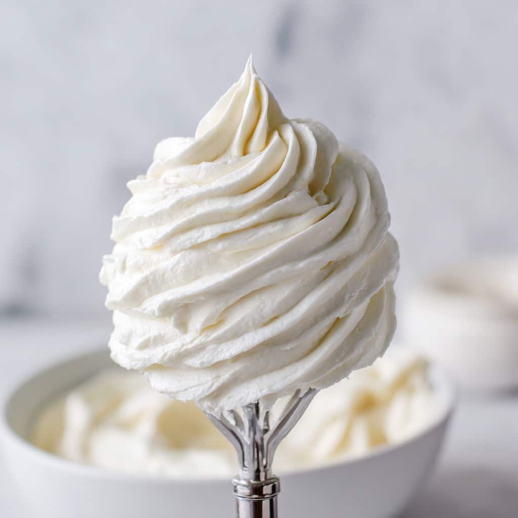 The image shows a close-up of a large, thick swirl of smooth, creamy white frosting held on the tip of a mixer paddle attachment. The frosting has a soft, fluffy texture with slight ridges and a few tiny air bubbles, suggesting it is whipped and light. In the blurred background, a white bowl filled with more of the same frosting can be seen, resting on a white marbled surface. The entire scene has a soft light that highlights the creamy, glossy finish of the frosting photo taken with an iphone --ar 4:5 --v 7