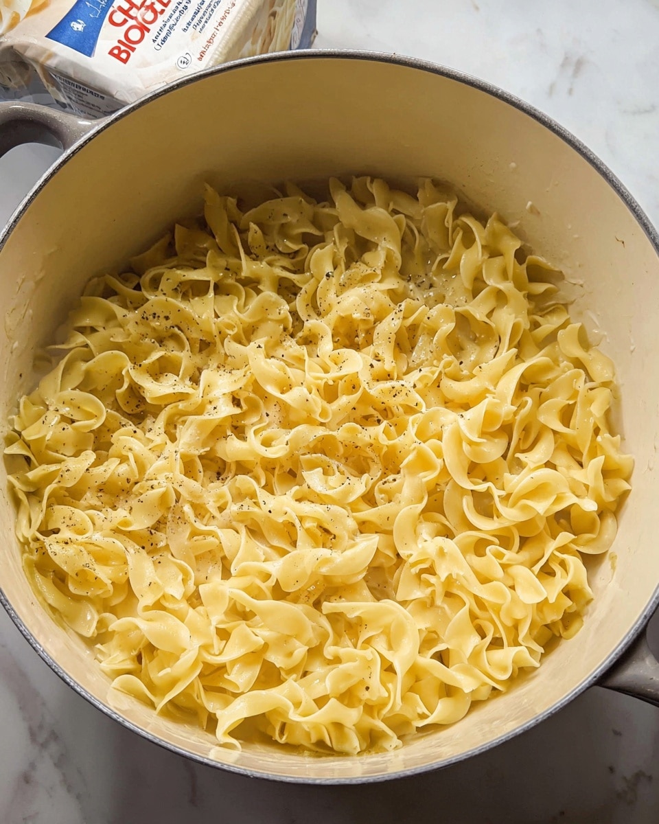 The image shows a close-up of cooked egg noodles with a glossy, creamy yellow sauce coating each curly pasta piece. The noodles are in a white pot with soft edges and a white spatula lifting a portion, highlighting the smooth texture and slight black pepper specks on the pasta. The creamy sauce gives the noodles a shiny, moist appearance, with some noodles curling loosely and overlapping each other. The background is a white marbled texture. Photo taken with an iphone --ar 4:5 --v 7