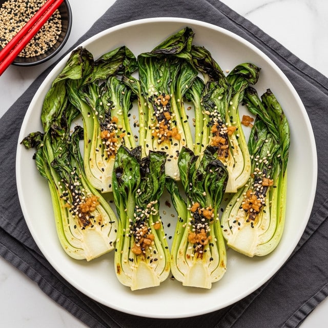 The dish shows six grilled baby bok choy halves arranged side by side in a white bowl, each piece with charred light golden brown edges and bright green leafy tops with some darkened parts. The bok choy is sprinkled with white and black sesame seeds and small bits of sautéed garlic or shallots. The bowl sits on a dark gray cloth over a white marbled surface. In the top left corner, a small dark bowl contains a mix of white and black sesame seeds, with red chopsticks resting beside it. Photo taken with an iphone --ar 4:5 --v 7