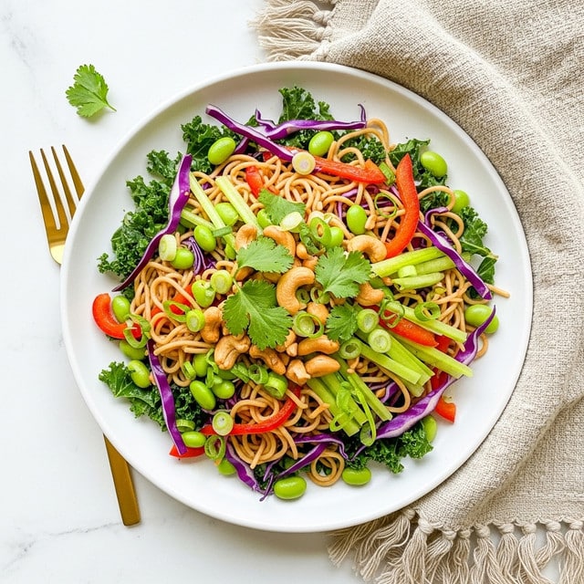The dish is a colorful noodle salad served on a white plate placed on a white marbled surface. The base layer is made of light brown cooked noodles, mixed with thin strips of dark green kale and purple cabbage. Scattered throughout are bright green edamame beans, thin slices of light green celery, and red bell pepper pieces adding brightness. There are also small golden-brown cashews on top, garnished with fresh cilantro leaves and thinly sliced green onions, giving the dish a fresh and crunchy look. A gold fork is placed next to the plate, and a beige textured cloth with fringes is nearby. Photo taken with an iphone --ar 4:5 --v 7