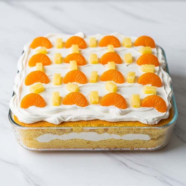 The image shows a square dessert in a clear glass dish with two layers: the bottom layer is a thick, moist yellow cake with a soft, crumbly texture, and the top layer is a thick, white whipped cream spread evenly across the cake. On top of the whipped cream layer, there are whole bright orange mandarin orange slices and small pale yellow pineapple chunks arranged evenly in rows, adding a colorful and fresh look. The background is a white marbled surface. Photo taken with an iphone --ar 4:5 --v 7
