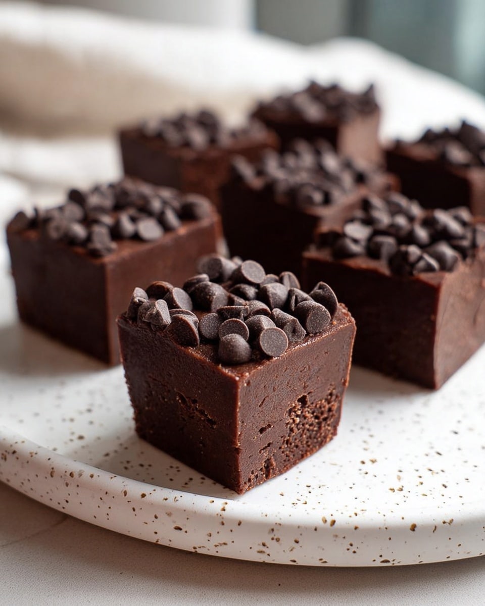 The image shows four dark chocolate fudge squares arranged on a white plate with small black speckles. Each square has a smooth, dense texture with visible vertical layers inside, showing a rich, creamy chocolate filling. The two front squares have bites taken out, revealing the thick and shiny inside layers, while the top square sits angled on the front one. Tiny dark chocolate chips cover the top of each square, adding a grainy texture contrast. The background is a white marbled surface with a soft, warm light in the scene. photo taken with an iphone --ar 4:5 --v 7