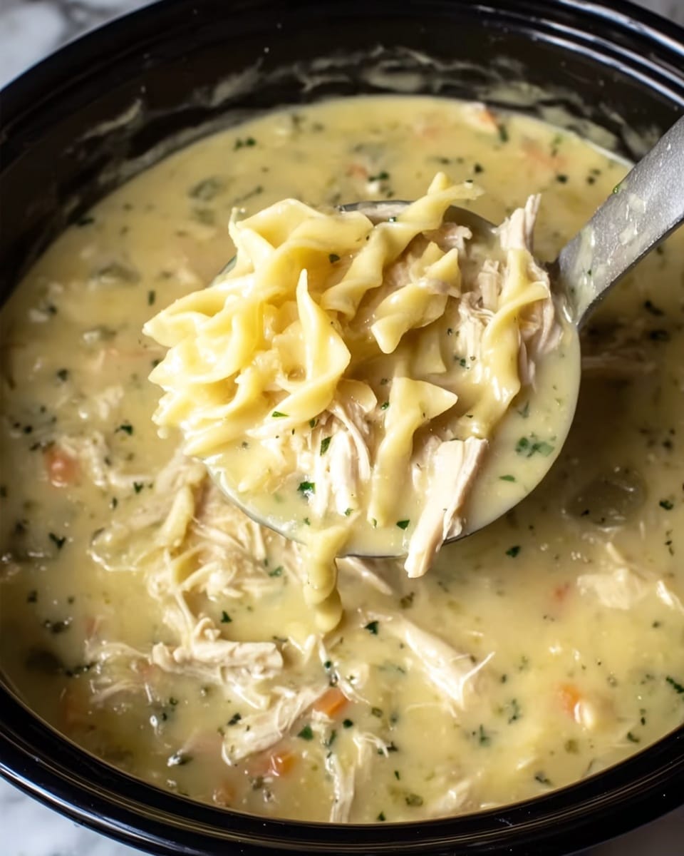 A white cup filled with thick, creamy chicken noodle soup sits on a white plate with a shiny spoon beside it. The soup includes pale yellow broth, short white noodle pieces, and chunks of light brown chicken throughout. Two halves of a biscuit, topped with melted butter, rest on the plate in front of the cup. A blue cloth with a white pattern is placed behind the cup, and the whole scene is on a white marbled surface. Photo taken with an iphone --ar 4:5 --v 7