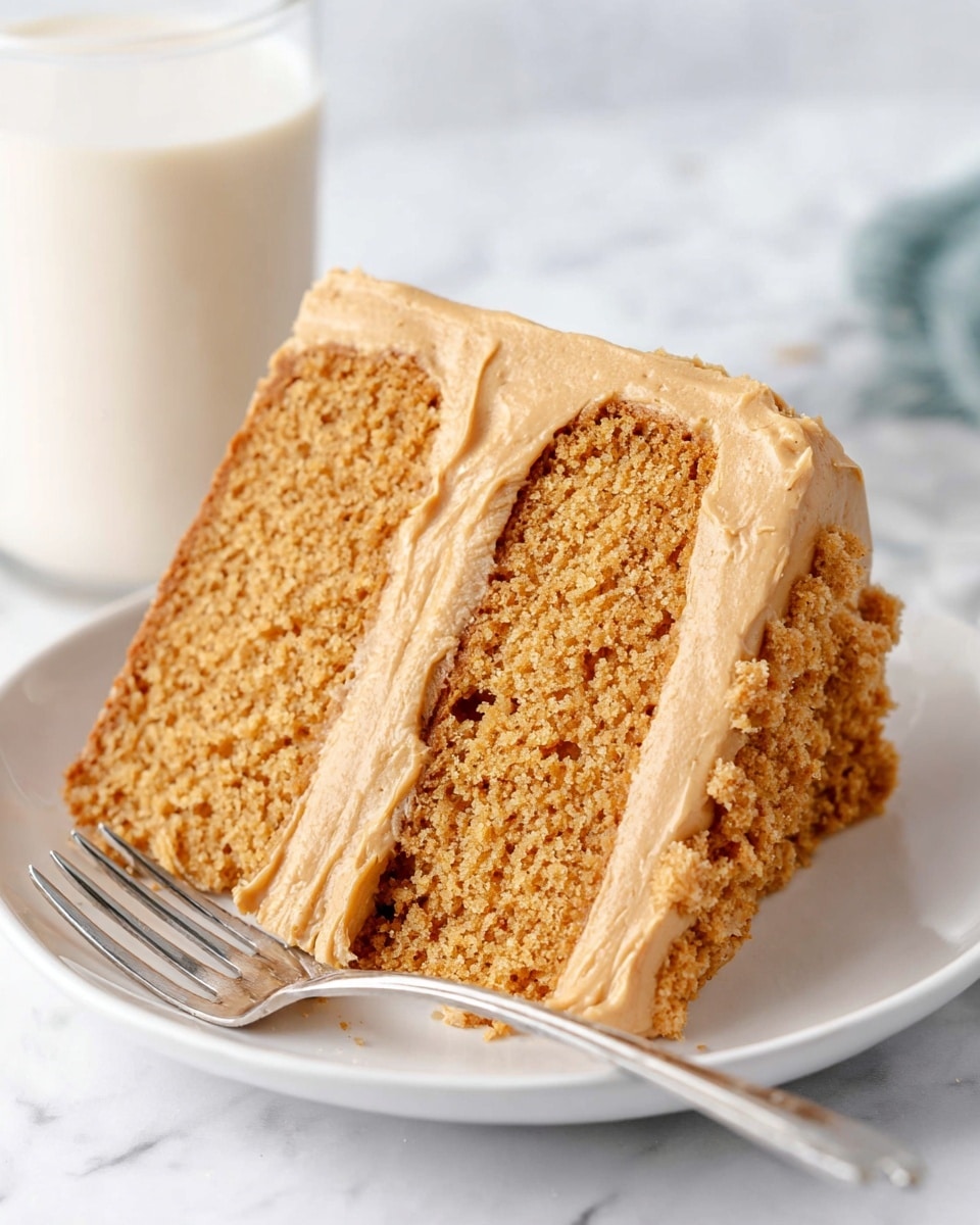 A round cake with a thick layer of light brown frosting evenly spread around its sides, showing visible smooth swirls and texture. The cake has crumbled brown cookie pieces scattered on top as decoration. It is placed on a white plate against a white marbled texture background. Photo taken with an iphone --ar 4:5 --v 7