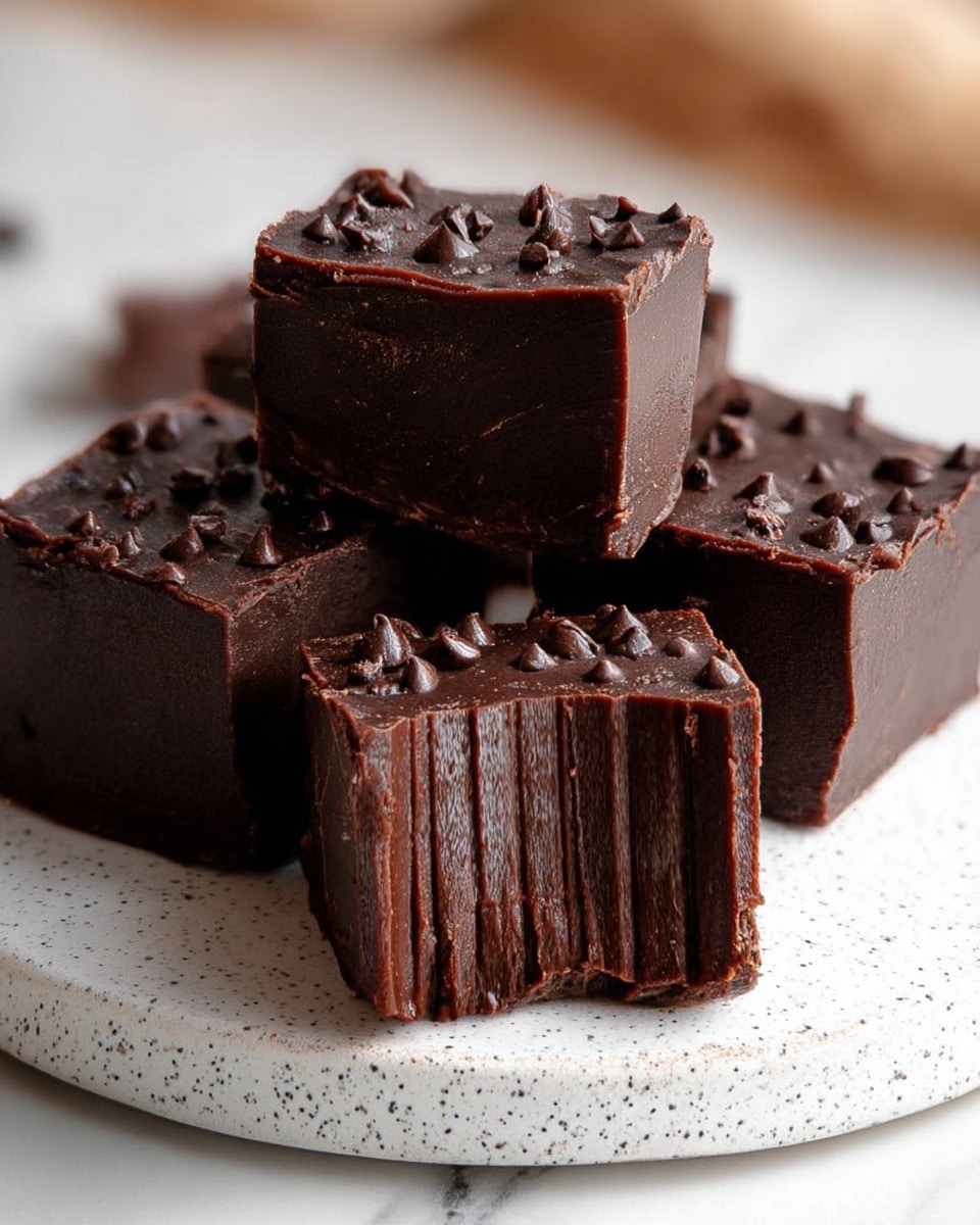The image shows square-shaped dark chocolate fudge cubes placed on a white speckled plate with a white marbled background. Each fudge cube has a dense, rich dark brown texture with visible small air holes and is topped with a thick layer of mini dark chocolate chips, giving a textured and slightly glossy look. The cubes are arranged in a scattered group, with some in focus and others blurred in the background. The edges of the fudge are sharp and clean, emphasizing their firmness. Photo taken with an iphone --ar 4:5 --v 7