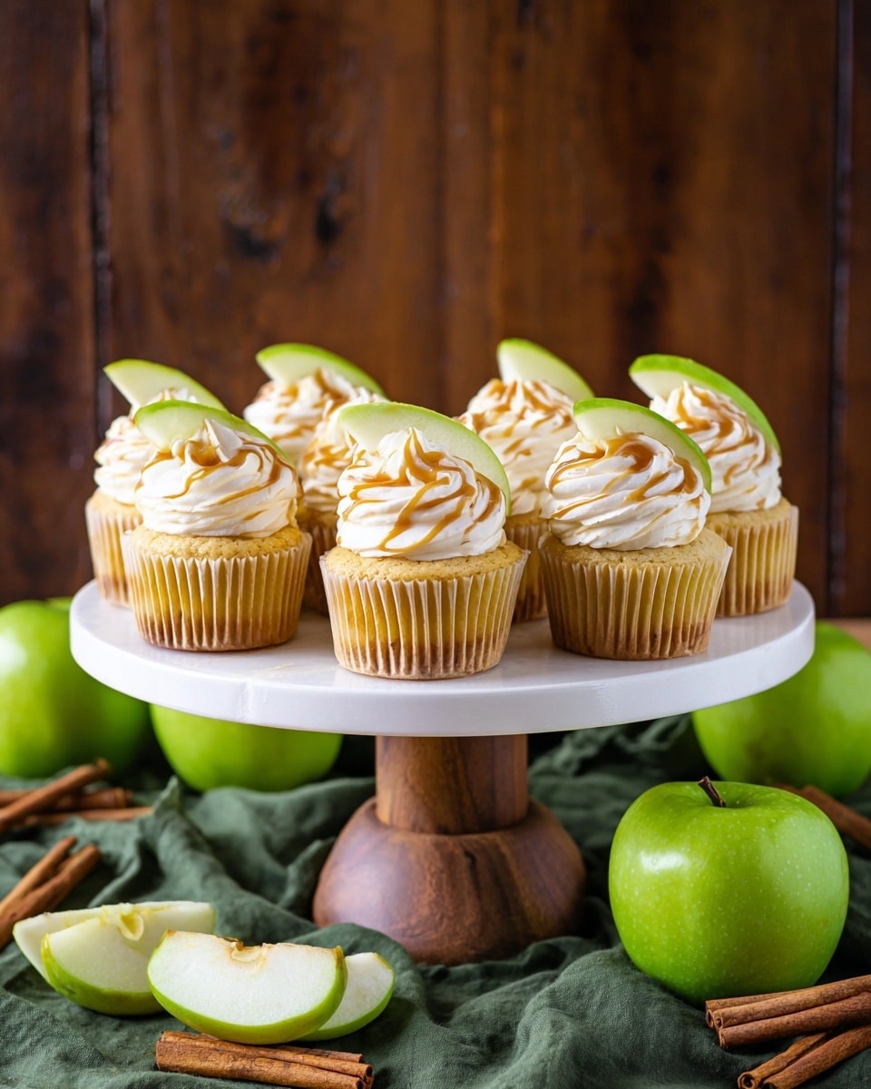 Nine cupcakes are displayed on a white round cake stand with a wooden base, each cupcake has a golden-brown base and a swirl of creamy off-white frosting on top. There is a thin drizzle of caramel sauce over the frosting. Each cupcake is topped with a thin, bright green slice of apple, partially inserted into the frosting. The scene includes bright green whole apples and apple slices around the base of the stand on a folded green cloth, all set on a white marbled surface. Photo taken with an iphone --ar 4:5 --v 7