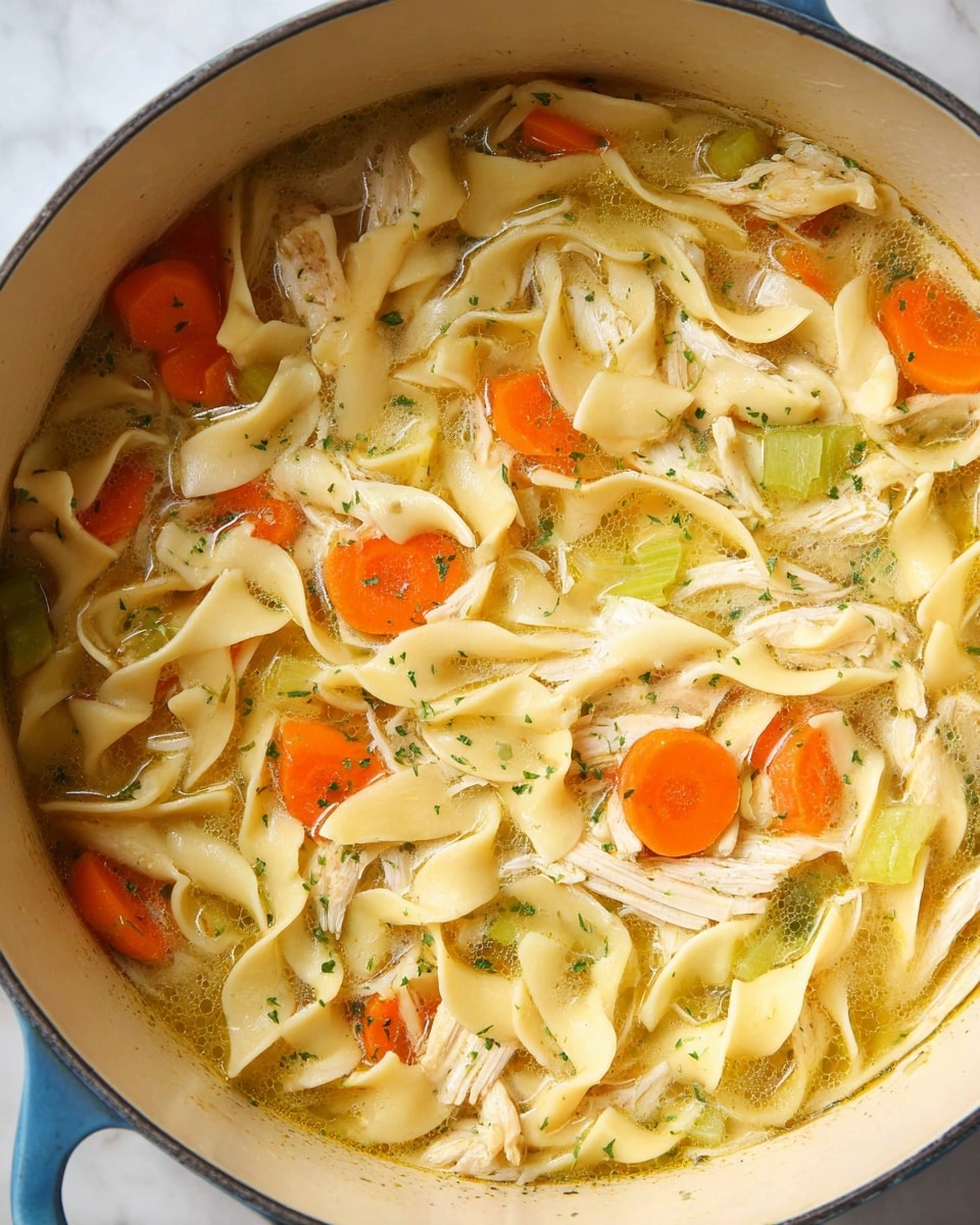 The image shows a close-up of a white pot filled with chicken noodle soup. The soup has broad, flat noodles that are pale yellow and soft, scattered throughout the pot along with thick slices of bright orange carrot rounds and small chunks of light green celery. Shredded pieces of cooked chicken with a light beige color are mixed in layers with the noodles and vegetables. The broth is a warm golden color with a slightly oily surface and is sprinkled with small green herb flakes. The pot sits on a smooth white marbled surface. photo taken with an iphone --ar 4:5 --v 7