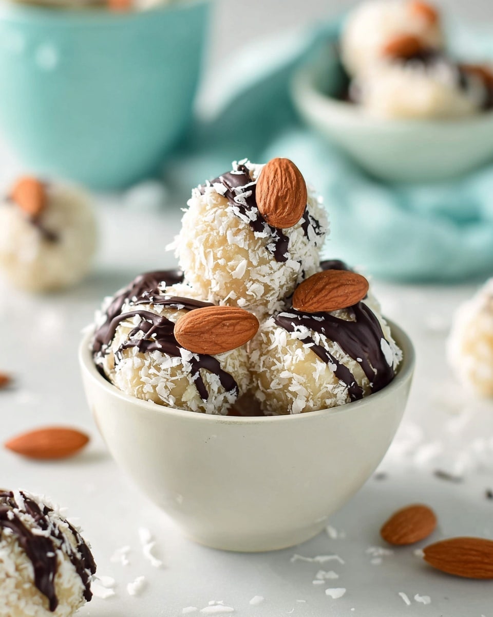 A small white bowl filled with round balls covered in white shredded coconut, each topped with a drizzle of dark chocolate and a single whole almond stuck in the chocolate layer near the top; the balls have a rough texture from the coconut flakes, and a few more almond-covered coconut balls are scattered on a white marbled surface around the bowl, creating a light and clean setting, with some blurred background elements and a pale blue bowl slightly out of focus behind. photo taken with an iphone --ar 4:5 --v 7