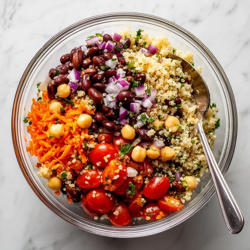 A clear glass bowl holds a fresh salad with several layers of ingredients visible: a base of light beige quinoa grains mixed with light yellow chickpeas, dark brown black beans, bright red halved cherry tomatoes, finely chopped orange carrots, bits of purple red onion, and small green herb pieces sprinkled throughout. A silver spoon is inside the bowl on the right side. The bowl is placed on a white marbled surface, making the colors of the salad stand out clearly. photo taken with an iphone --ar 4:5 --v 7
