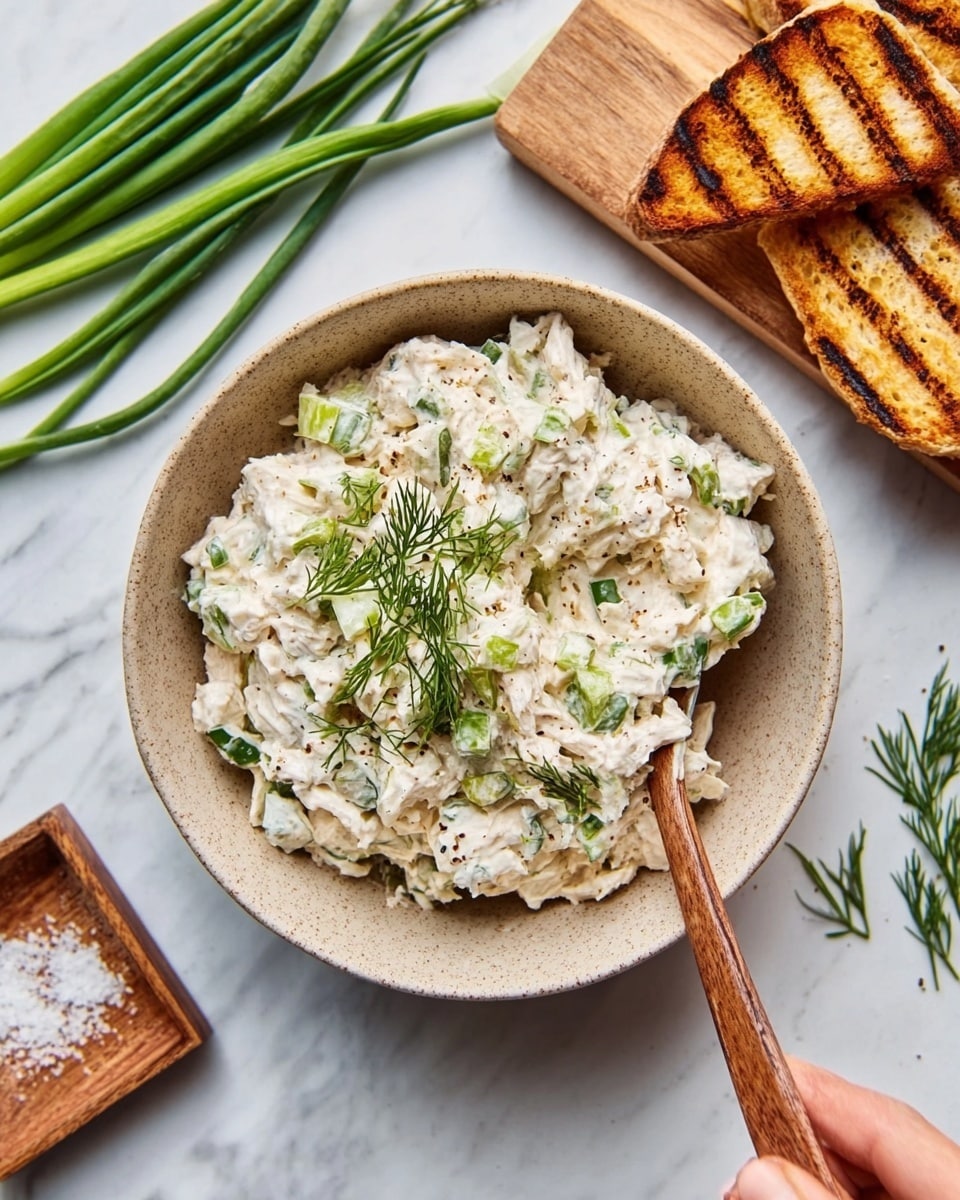 A white bowl filled with a creamy, chunky tuna salad mixed with small green herbs sits centered on a white plate. The tuna salad layer is textured with bits of tuna and herbs, appearing moist and thick. Around the bowl, uneven pieces of pale beige crackers are arranged, contrasting with the smoothness of the salad. A wedge of light green avocado leans against the tuna salad inside the bowl. The setting rests on a white marbled surface. photo taken with an iphone --ar 4:5 --v 7