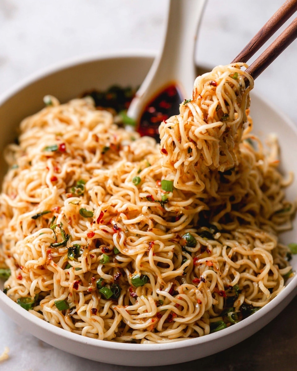 A close-up of a bowl filled with cooked noodles mixed with small green chopped onions and specks of red chili flakes, giving the dish a textured look with colors of light yellow, green, and red. The noodles appear glossy and slightly oily, neatly held by wooden chopsticks lifting a portion from the bowl, which is white. To the side inside the bowl, a white ceramic spoon rests with some dark red and green sauce and oil. The background surface is a white marbled texture. photo taken with an iphone --ar 4:5 --v 7