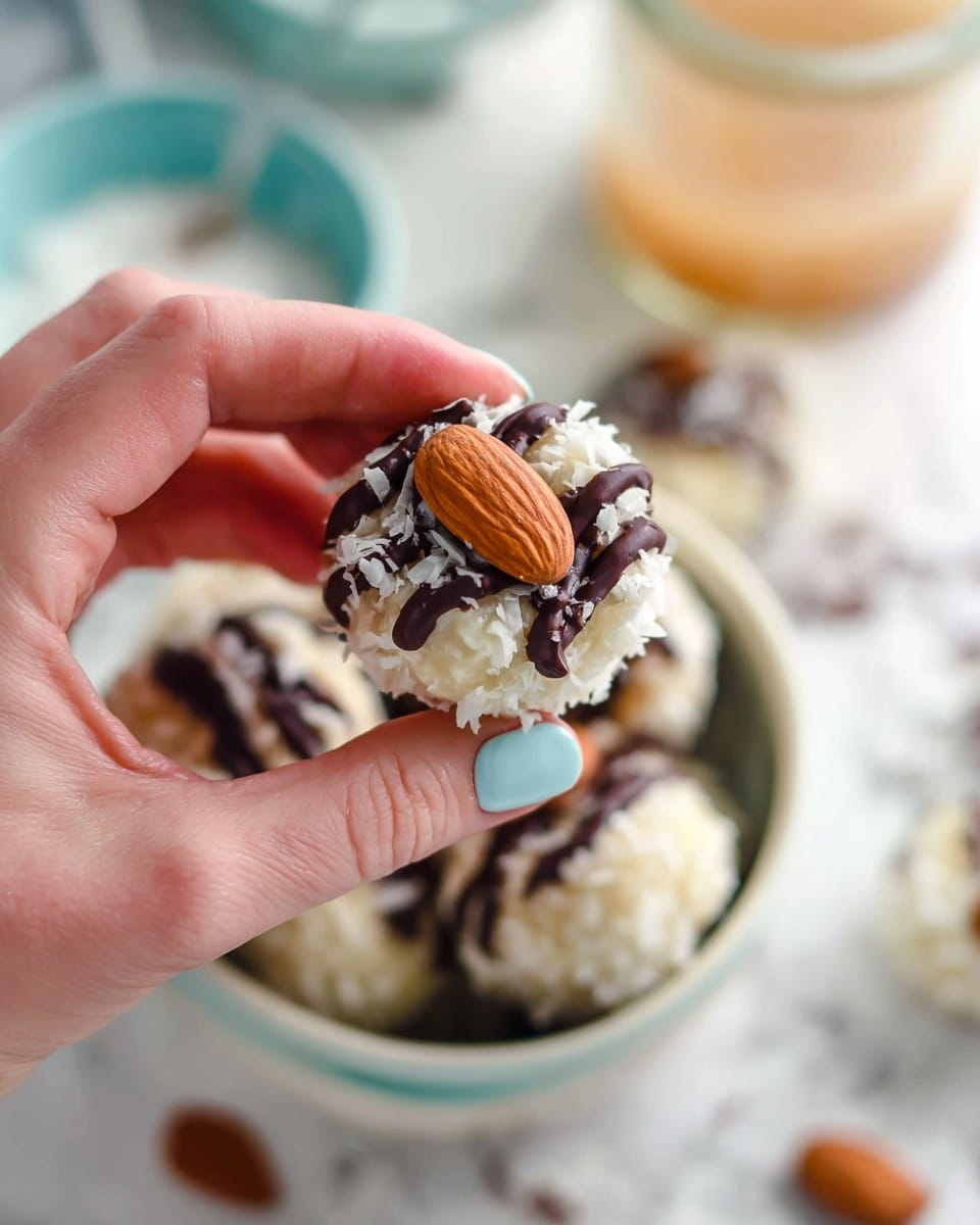 A close-up view of a round sweet treat held by a woman's hand with painted light blue nails, showing three main layers: the bottom soft white layer coated in shredded coconut, a drizzle of dark chocolate on top creating stripes, and a whole almond placed in the center. In the background, several similar treats sit inside a white bowl with a light blue exterior, all placed on a white marbled surface. Blurry objects like a small glass jar with a light colored drink and a blue bowl are faintly visible. photo taken with an iphone --ar 4:5 --v 7