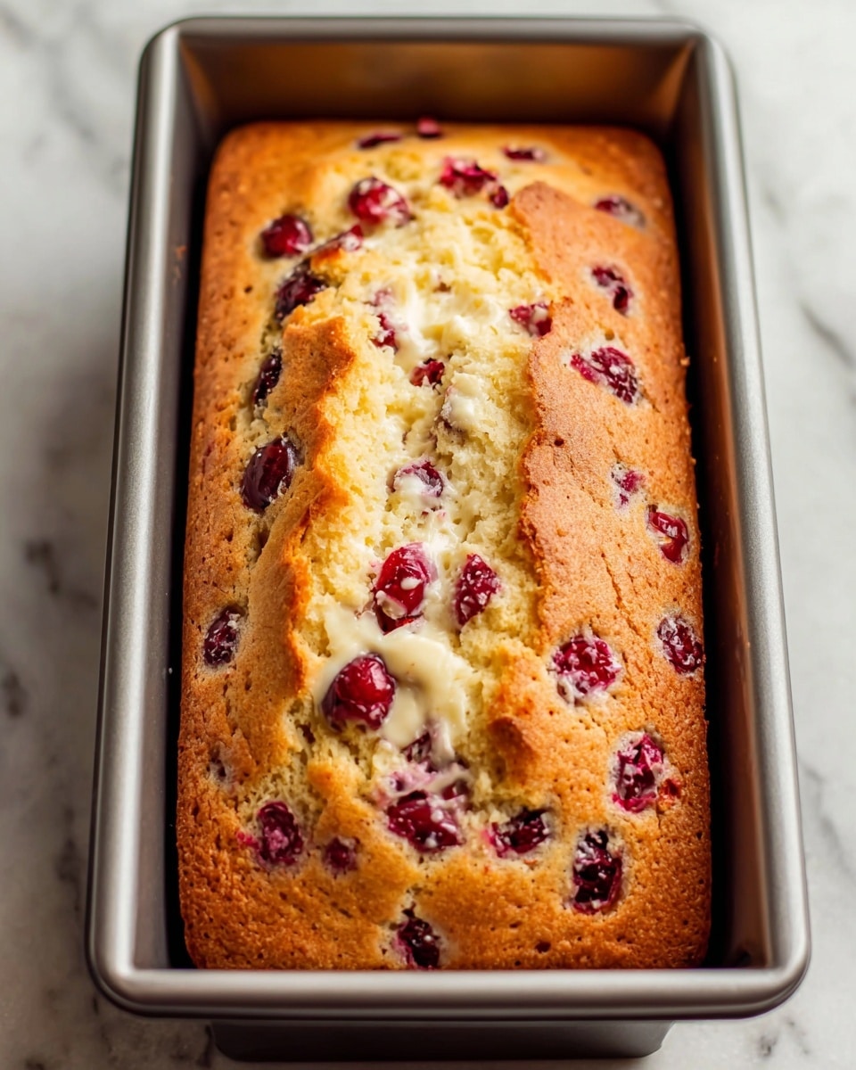 A freshly baked rectangular loaf cake sits in a silver baking pan on a white marbled surface, with a soft, golden-brown crust and a slightly cracked top. The cake is embedded with whole and halved red cranberries, adding bright pops of deep red throughout the light, fluffy texture. Creamy white patches are visible where melting white chocolate or cream cheese is mixed within the cake, creating a moist and rich appearance. The edges are slightly darker and firm, contrasting with the inviting, tender interior. Photo taken with an iphone --ar 4:5 --v 7