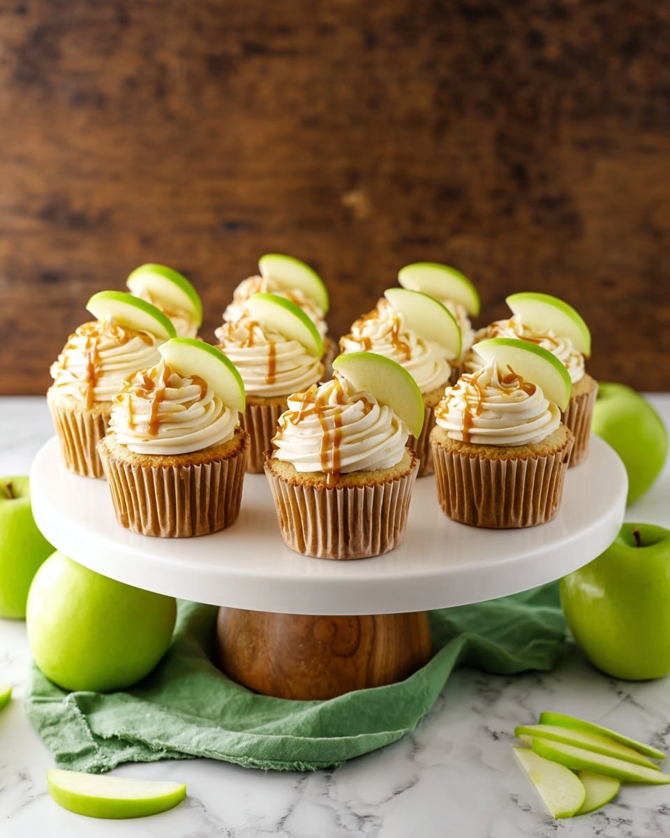 A white cake stand with a wooden base holds nine cupcakes arranged in three rows of three. Each cupcake has a golden-yellow base with a soft texture, topped with a large swirl of smooth white cream frosting. On top of the frosting, there is a light caramel drizzle and a thin green apple slice inserted at the back edge, adding a fresh green color. The stand is placed on a wooden table covered with a green cloth, and around it are whole and sliced green apples and cinnamon sticks. The background is a warm dark wood, and the whole scene is lit softly, making the cupcakes look fresh and inviting. Photo taken with an iphone --ar 4:5 --v 7