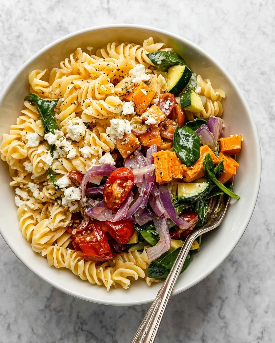 A white bowl holds a colorful pasta salad with three main layers: at the bottom is a bed of light yellow, curly pasta spirals, middle layer has bright green spinach leaves, chunks of dark green zucchini, orange sweet potatoes, and slices of purple onion mixed throughout, and the top layer is scattered with small pieces of crumbled white cheese and halved red cherry tomatoes, with bits of black pepper seasoning visible; a silver fork rests on the right edge of the bowl, all set on a white marbled surface. photo taken with an iphone --ar 4:5 --v 7