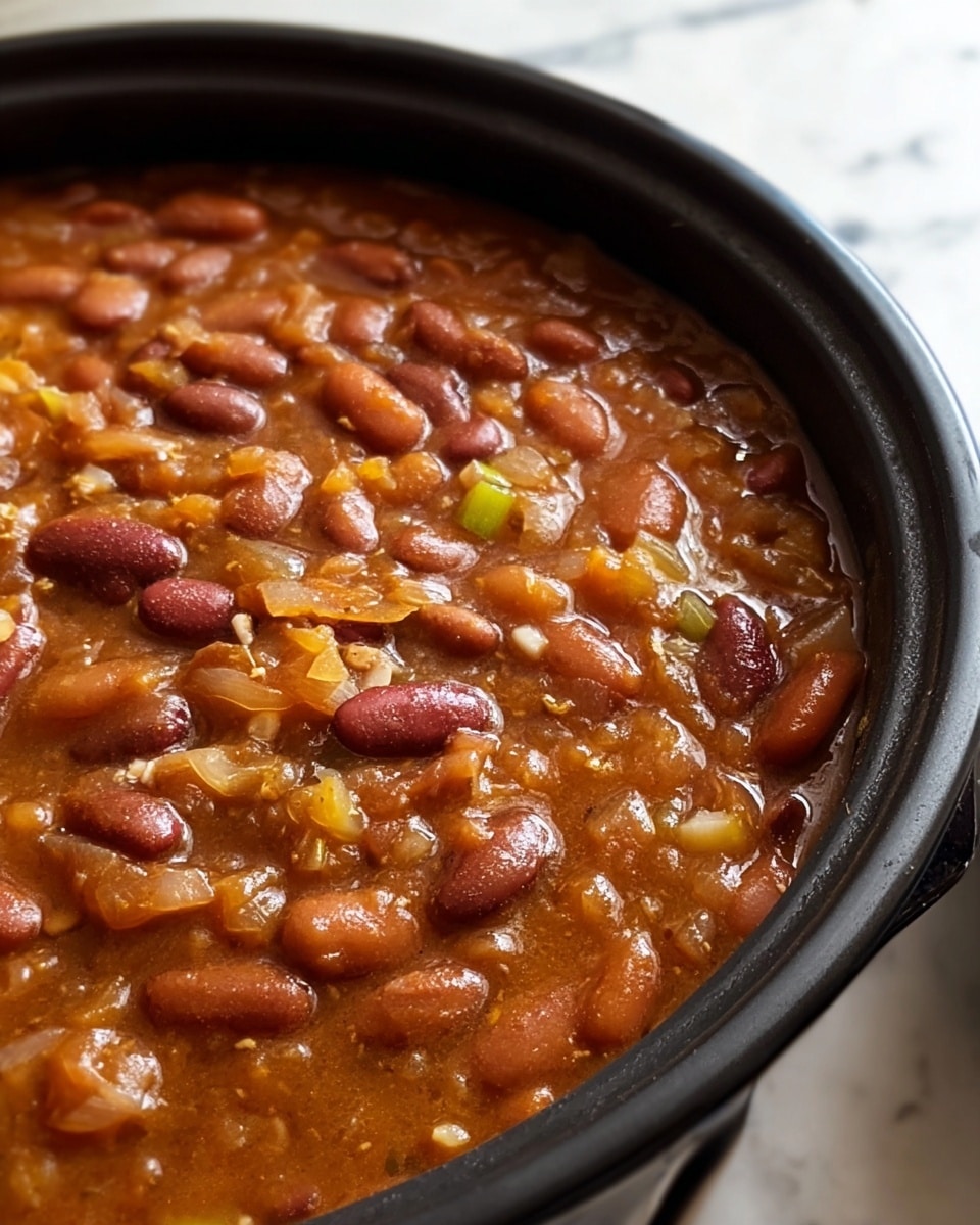 The image shows a close-up of a black pot filled with a thick stew. The stew has a brownish-orange broth filled with many beans, appearing in light beige and dark red colors. There are visible pieces of translucent onion and small chunks of yellow bell pepper mixed in. The stew looks textured and hearty, with bits of ground meat or mashed beans adding to its rich, slightly chunky consistency. The pot sits on a white marbled surface with some fabric draped in the background. photo taken with an iphone --ar 4:5 --v 7