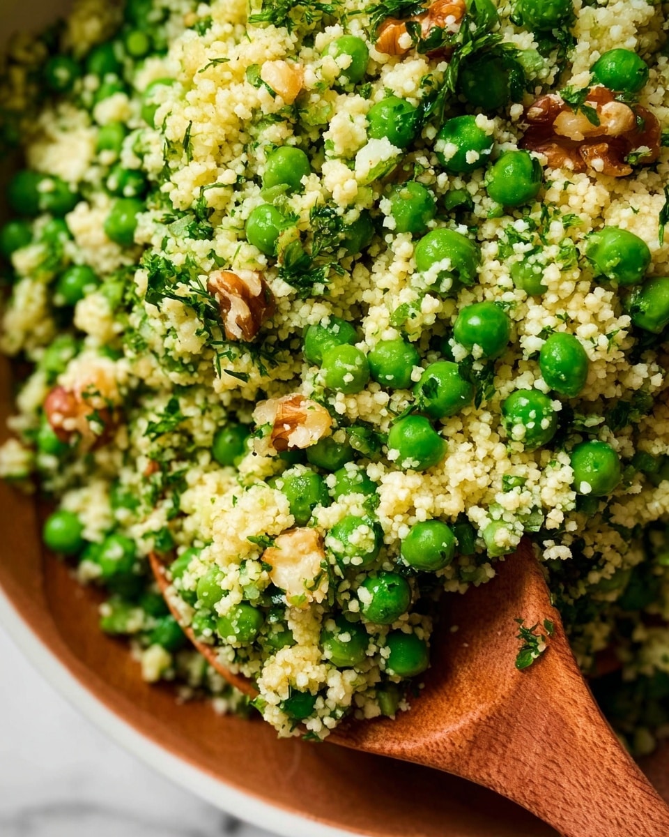This close-up image shows a bowl filled with a green and white mixed salad. The main layer is small, light yellow couscous grains scattered throughout, with bright green peas mixed in evenly. Small pieces of chopped green herbs are sprinkled across the dish, adding fresh color. Some light brown walnut pieces are also visible, adding texture and contrast. A wooden spoon is partially submerged in the salad, showing some couscous grains sticking to it. The bowl itself is white, and the background has a white marbled surface texture. Photo taken with an iphone --ar 4:5 --v 7