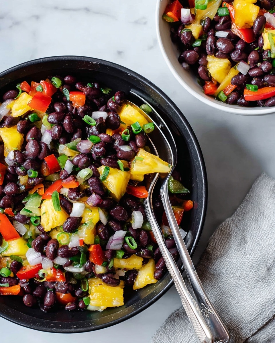 A white bowl filled with a colorful black bean salad sits on a white marbled surface, resting partially on a light gray cloth. The salad has three main layers: the bottom layer consists mostly of black beans scattered throughout, the middle layer shows chunks of red bell pepper and yellow pineapple pieces mixed evenly, and the top has sliced green onions and small rings of white onion sprinkled over. Two silver spoons lie inside the bowl, ready for serving. The colors are bright and fresh, making the dish look vibrant and appetizing. Photo taken with an iphone --ar 4:5 --v 7