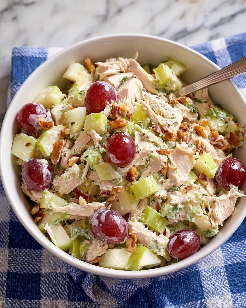 A white bowl filled with a mixed salad, showing several layers including light green celery slices, pale pink shredded chicken, and halved red grapes scattered throughout. There are also small pieces of chopped walnuts sprinkled on top, adding a crunchy texture. The salad looks creamy with a white dressing coating the ingredients, and some light green herbs are dotted across the surface. A silver spoon with an ornate handle is placed inside the bowl, and the bowl sits on a white marbled surface partially covered by a blue and white checkered cloth. photo taken with an iphone --ar 4:5 --v 7