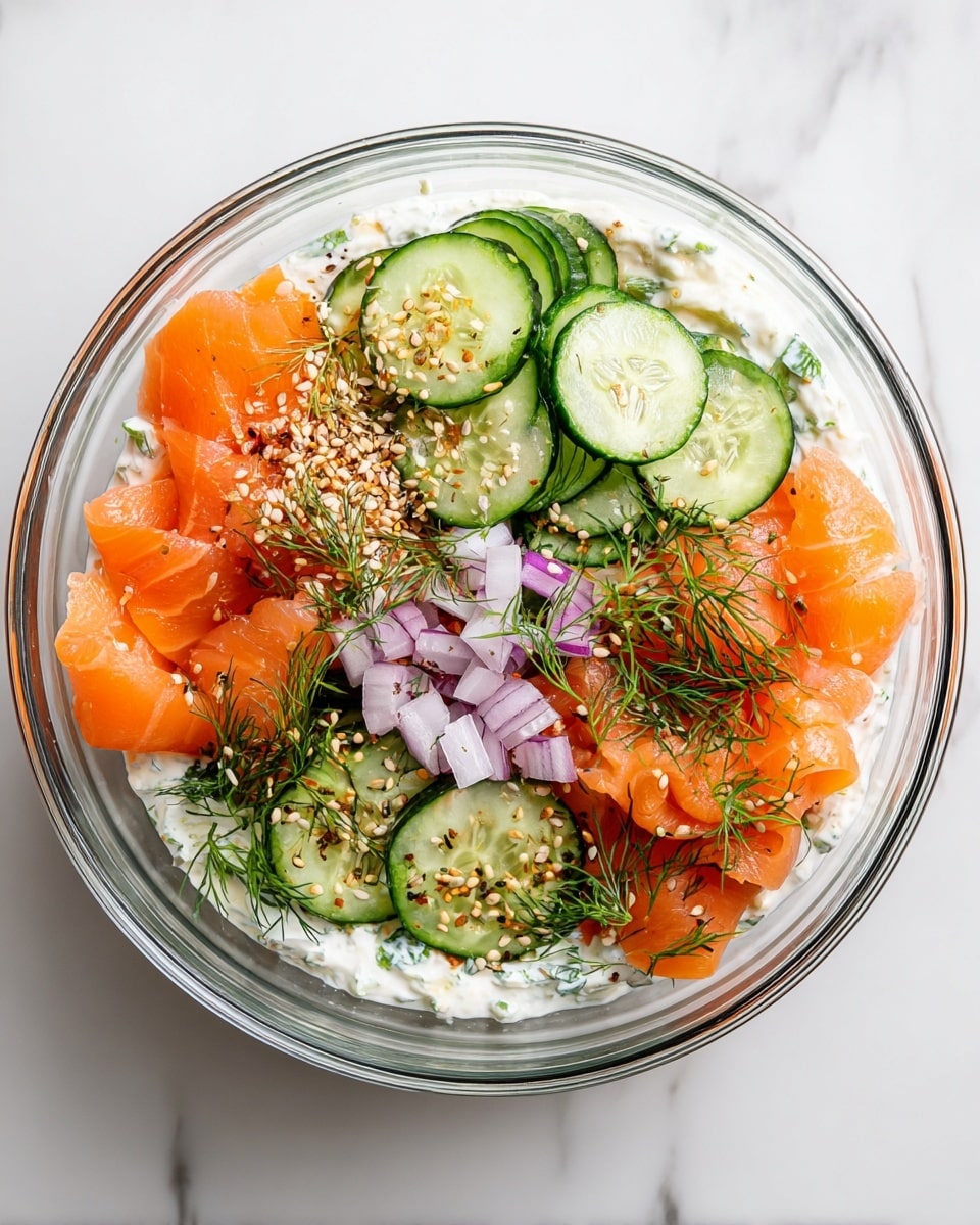 A clear glass bowl holds this dish with three layers visible from above. The bottom layer is white and creamy, covering the entire base. On top, there are thin slices of bright orange smoked salmon placed on the left side, slightly folded and sprinkled with small green dill leaves and a mix of white, black, and orange seeds. To the right of the salmon, there are slices of fresh green cucumber, arranged loosely with some small pieces of purple onion scattered in between, all topped with the same seed and herb mix. The bowl sits on a white marbled surface. photo taken with an iphone --ar 4:5 --v 7