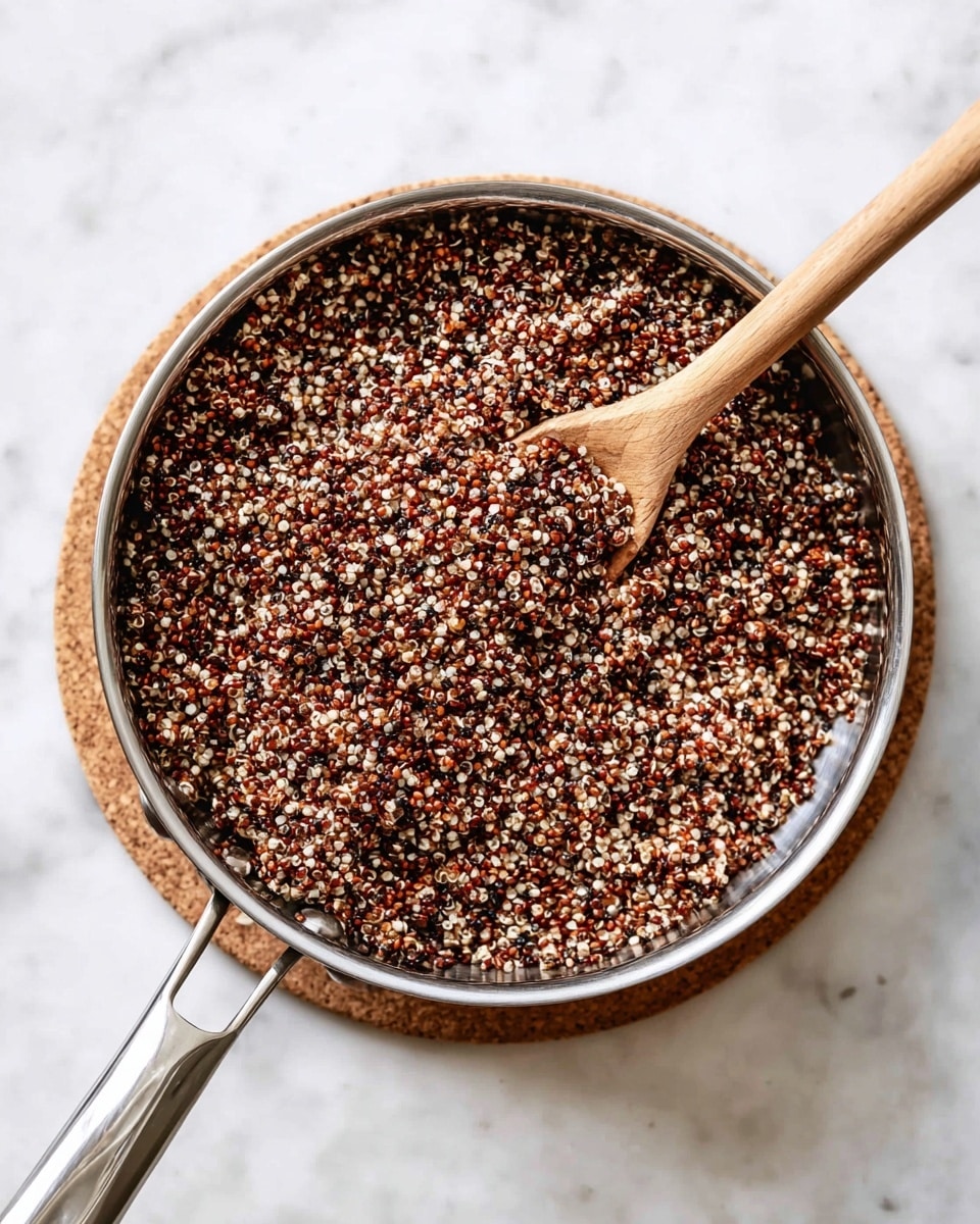 A close-up top view of a metal pan filled with cooked tri-color quinoa, showing layers of small round grains in shades of brown, red, and white all mixed together with a slightly fluffy texture. A light wooden spoon with a long handle is resting inside the pan, partly covered by the quinoa. The pan is placed on a round cork mat, and the whole scene is set on a white marbled surface. photo taken with an iphone --ar 4:5 --v 7
