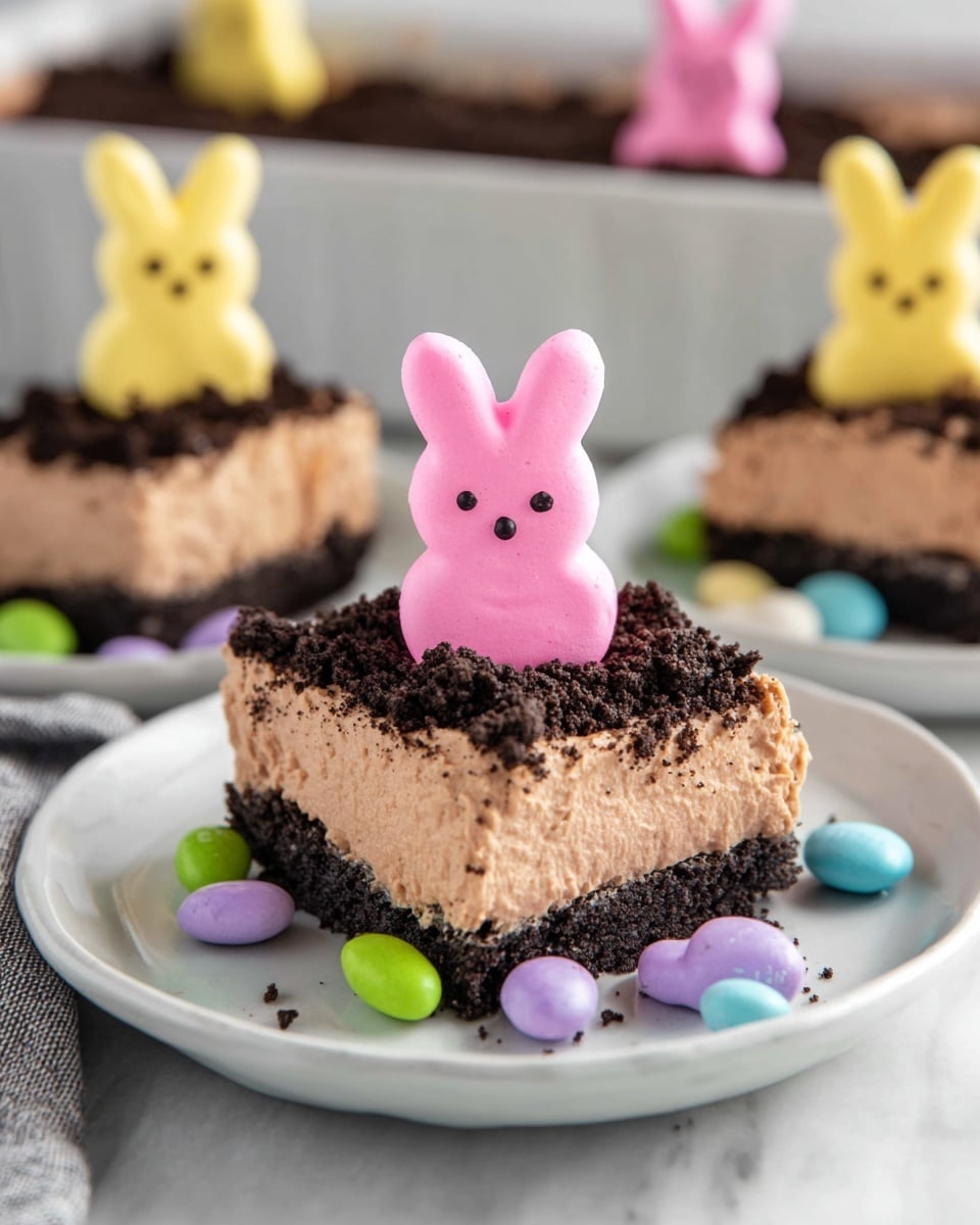 A square dessert slice is served on a white plate set on a white marbled surface. The dessert has three main layers: a dark, crumbly cookie crust on the bottom, a thick middle layer of smooth, light brown chocolate mousse, and a top layer of crushed dark cookies. On top, a bright pink marshmallow bunny stands upright, decorated with small dark chocolate dots for eyes and nose. Around the dessert, scattered colorful candy-coated chocolates in pastel shades of purple, green, blue, and yellow add a cheerful touch. In the background, part of a white baking dish with more dessert and additional yellow and purple marshmallow bunnies is visible. Photo taken with an iphone --ar 4:5 --v 7