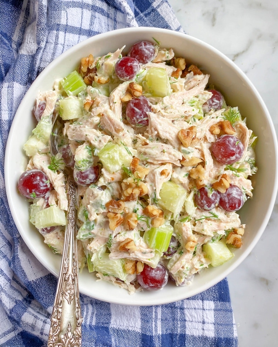 A white bowl filled with a mixed salad that has several visible layers and textures. The base layer consists of chopped light green celery sticks and pale yellow apple slices cut into small pieces. Mixed throughout are pale pink shredded chicken pieces that look tender. Scattered on top are halved red grapes with a shiny surface, and small brown chopped nuts adding crunch. The salad is lightly coated in a creamy white dressing, with dark green herbs sprinkled over for color. The bowl is placed on a blue and white checkered cloth on a white marbled surface, and a silver spoon is partially visible inside the bowl. photo taken with an iphone --ar 4:5 --v 7