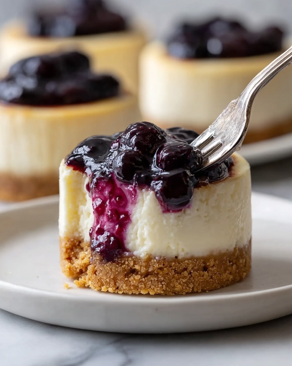 A close-up image of a small cheesecake with three visible layers, resting on a white plate on a white marbled surface. The base layer is a crumbly, golden brown crust with a coarse texture. The middle layer is a thick, creamy off-white cheesecake that looks smooth and soft, and a fork is pressing into its right side, making a bite. The top layer is a glossy, dark purple blueberry sauce with visible whole berries, covering the top and slightly dripping down the side. In the blurry background, two more identical cheesecakes are visible but out of focus. Photo taken with an iphone --ar 4:5 --v 7