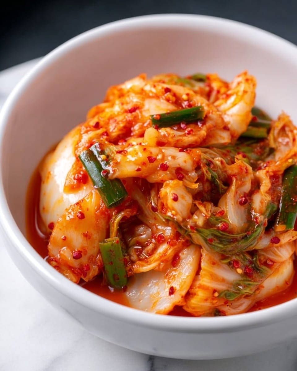 A white bowl filled with spicy kimchi showing multiple layers of reddish-orange fermented cabbage with visible red pepper flakes, light green onion pieces scattered throughout, and a glossy, wet texture. A pair of black chopsticks held by a woman's hand is lifting a portion of the kimchi, highlighting the wrinkled, soft cabbage leaves coated in sauce. The background is a white marbled texture. Photo taken with an iphone --ar 4:5 --v 7