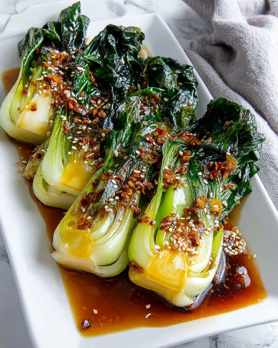 Four cooked baby bok choy are neatly lined up on a white rectangular plate, each one showing a clear layer of pale green and light yellow stems at the top, with dark green, wilted leafy ends at the bottom. The vegetables are glistening with a soy-based sauce that pools slightly on the plate, and scattered across them are small toasted sesame seeds and bits of red chili flakes. The plate is placed on a white marbled surface with a light gray cloth in the background, adding softness to the scene. A small square dish with sesame seeds is partly visible in the bottom left corner. photo taken with an iphone --ar 4:5 --v 7