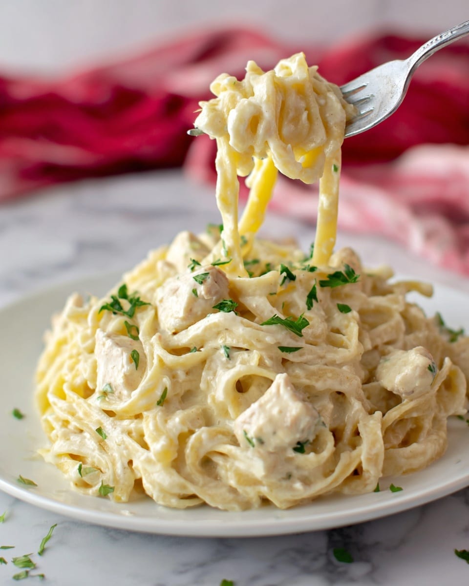 A close-up view of creamy chicken alfredo pasta served on a white plate, showing thick layers of pale yellow fettuccine noodles mixed with tender white chicken chunks covered in a rich, smooth, off-white cream sauce. Small bright green parsley pieces are scattered on top, adding color contrast. A silver fork holds a spoonful of the creamy pasta above the plate, with the noodles and sauce glistening. The plate is set on a white marbled surface with a red and white cloth in the blurred background. Photo taken with an iphone --ar 4:5 --v 7