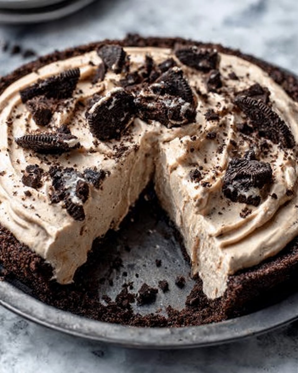 A round chocolate pie with a dark brown crumb crust forms the base layer, holding a thick, creamy light beige filling on top. The filling is smooth with swirls, spread evenly across the pie. Large pieces of broken chocolate cookies are scattered over the filling, adding dark, rough textured spots. The pie sits on a dark metal tray over a white marbled texture. A slice has been cut and slightly pulled out, showing the crisp crust and creamy filling clearly. Photo taken with an iphone --ar 4:5 --v 7