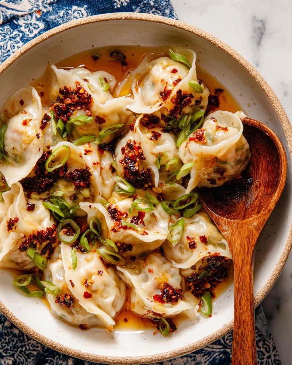 A close-up of a round white bowl filled with plump dumplings in a light, glossy sauce, each dumpling showing slightly translucent, soft, white wrappers with crinkled edges. Scattered on top are small pieces of dark red chili flakes and finely chopped bright green spring onions, adding texture and color contrast. A wooden spoon rests inside the bowl on the right side, partially submerged in the sauce, with a few dumplings overlapping it. The bowl sits on a white marbled surface with a blue and white patterned cloth partially visible underneath. photo taken with an iphone --ar 4:5 --v 7