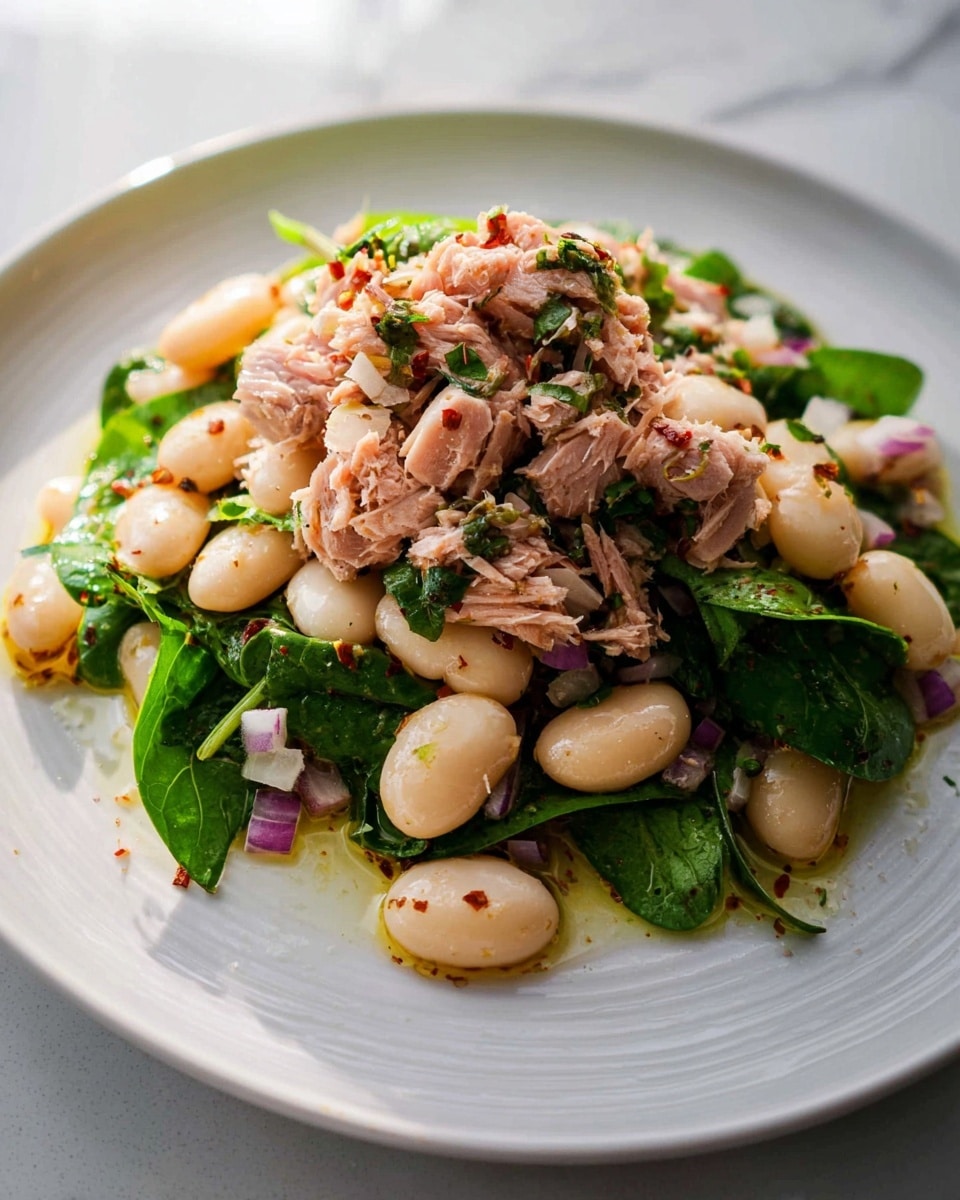 A close-up image of a white plate with a mixed salad of fluffy light pink shredded tuna on the top layer, combined with a bottom layer of large white beans, chopped green herbs, and small pieces of purple onion scattered throughout. The salad has a glistening, slightly oily texture that adds shine, and a silver fork rests beneath the salad with some beans and tuna on its surface. The background shows a blurred piece of toasted bread on a white marbled texture. photo taken with an iphone --ar 4:5 --v 7