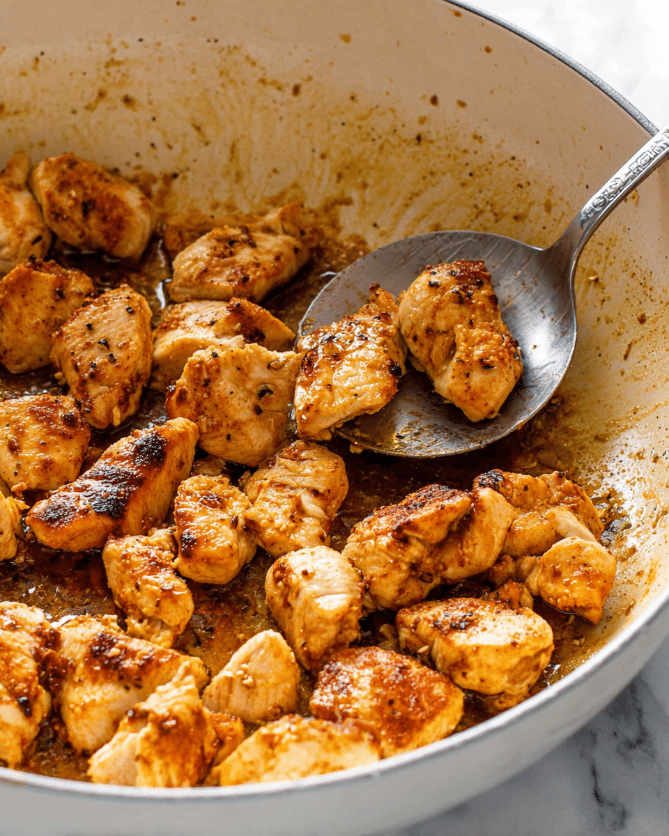 The dish is a single-layered mix of penne pasta and cooked chicken pieces in a white pot. The penne is golden-yellow and scattered evenly throughout. The chicken pieces are golden-brown with a slightly crispy texture on the edges, mixed well with small chunks of red and green bell peppers, and bits of tomato. The entire dish is coated lightly with a creamy orange-colored sauce, topped with small sprinkles of grated cheese and chopped green herbs. The pot sits on a white marbled surface with a wooden spoon resting nearby. Photo taken with an iphone --ar 4:5 --v 7