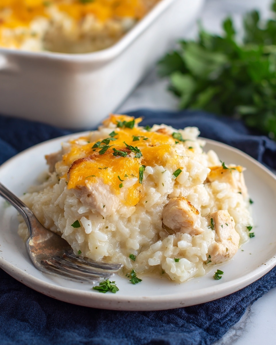 A serving of a creamy, white rice and chicken casserole sits on a white plate over a white marbled surface with a dark blue cloth underneath. The dish has one main thick layer of tender chicken pieces mixed into soft, moist white rice, topped with melted golden yellow cheese. Small green herb bits are scattered on top and around for color contrast. A silver fork rests partly on the plate’s edge near the food. In the blurred background, part of the casserole dish is visible along with more green herbs. Photo taken with an iphone --ar 4:5 --v 7