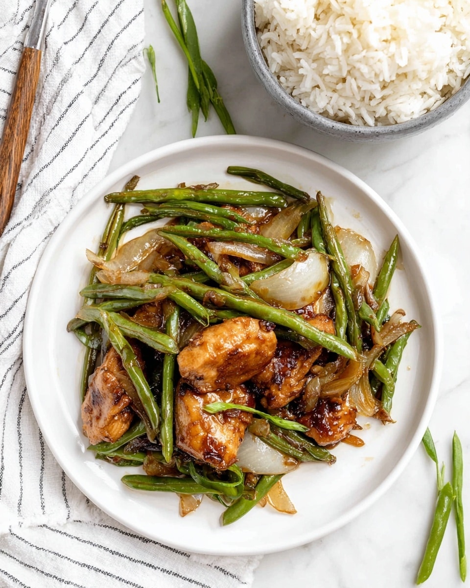 A white plate holds a stir-fry with several layers: at the bottom are long, green beans with a shiny, slightly wrinkled texture, mixed with translucent, lightly browned onion slices. On top are pieces of browned chicken with a glossy, caramelized surface and a rich golden color. Around the plate, fresh green beans are scattered. Above and slightly to the side, a gray bowl contains fluffy white rice with visible separate grains. The scene is set on a white marbled surface with a striped white cloth partially visible. Photo taken with an iphone --ar 4:5 --v 7