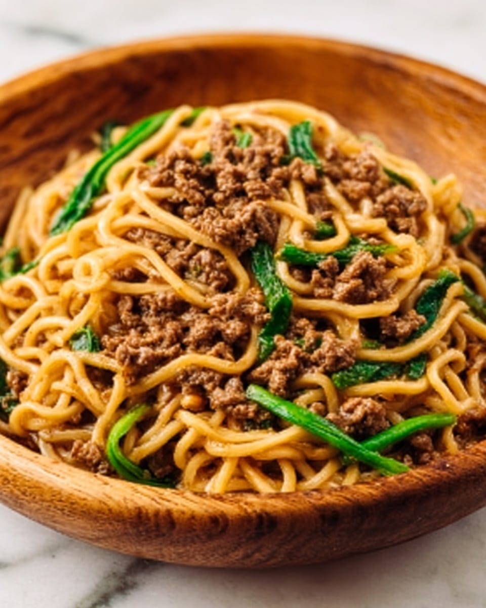 The image shows a close-up of a wooden bowl filled with noodles, mixed with ground meat and green vegetable strips. The noodles are light golden, thick, and covered with a sauce that gives them a slight shine. The ground meat is brown and crumbled, resting mostly on top and mingled throughout the noodles. Bright green vegetable strips are scattered evenly among the noodles and meat, adding color and freshness. The bowl sits on a white marbled surface. photo taken with an iphone --ar 4:5 --v 7